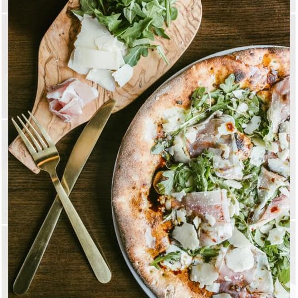 A pizza with prosciutto, arugula, and shaved parmesan, next to a board with more toppings and cutlery.