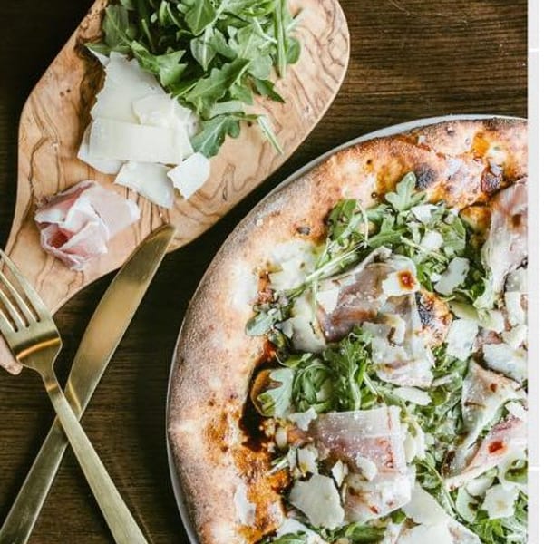 A pizza topped with arugula, prosciutto, and shaved parmesan, next to a wooden board with fresh ingredients.