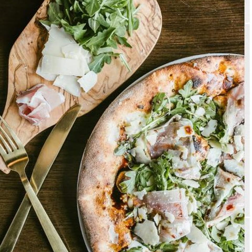A pizza topped with arugula, prosciutto, and shaved parmesan, next to a wooden board with fresh ingredients.