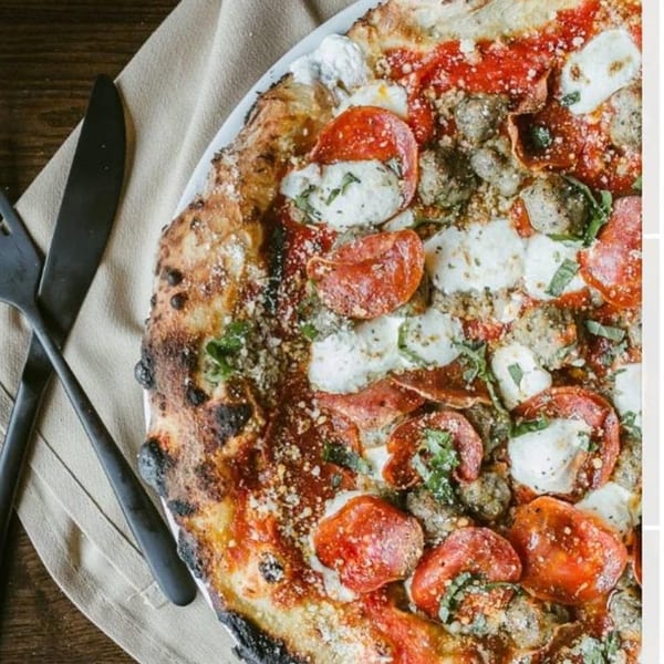 Close-up of a wood-fired pizza with pepperoni, sausage, mozzarella, and basil, next to cutlery.