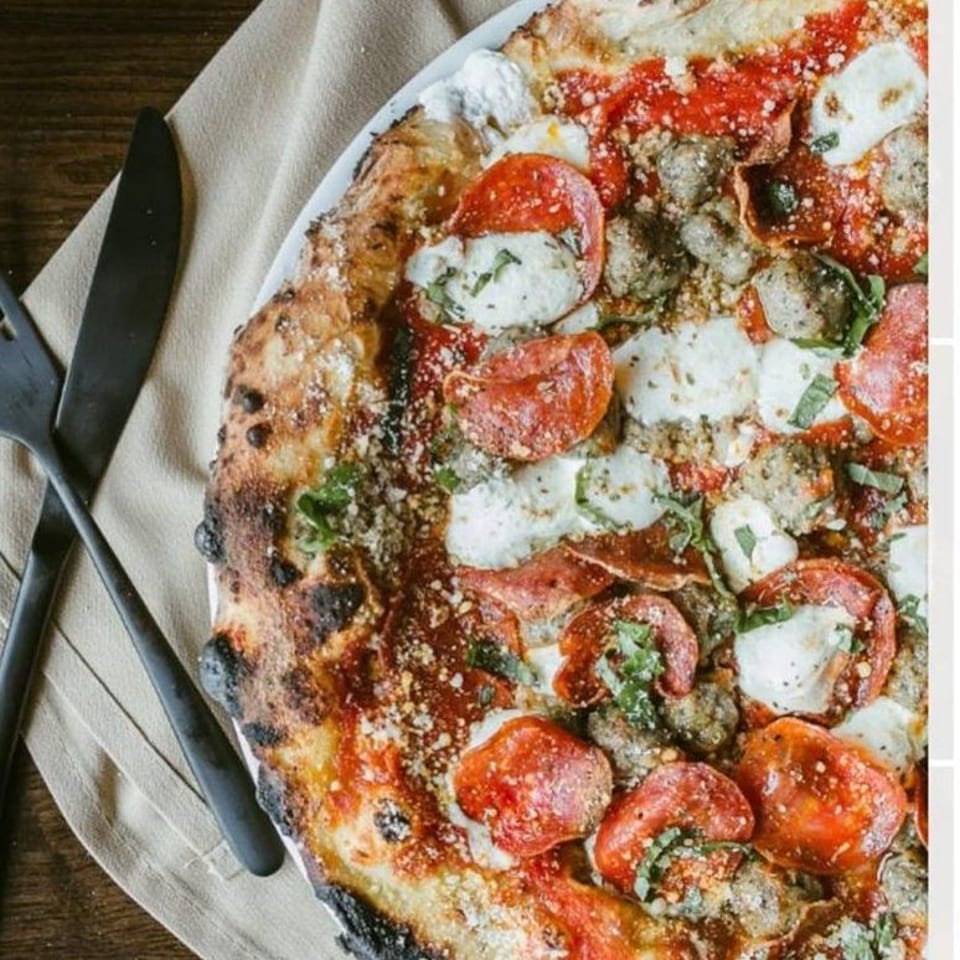 Close-up of a wood-fired pizza with pepperoni, sausage, mozzarella, and basil, next to cutlery.
